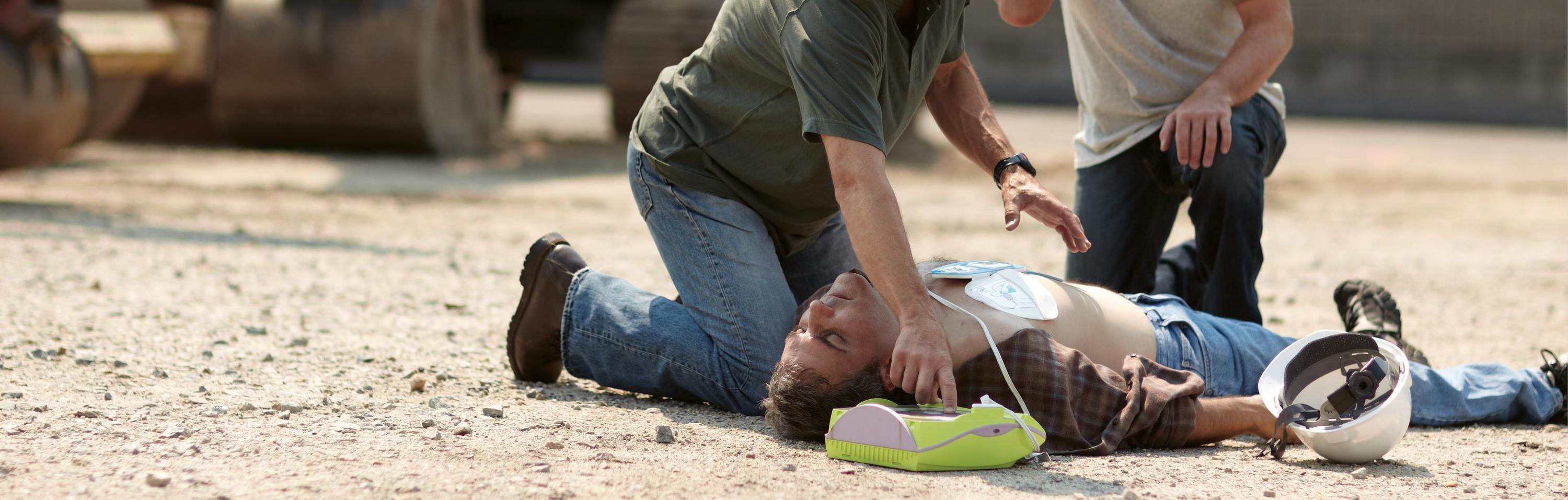 Ein Bauarbeiter erhält eine Wiederbelebungsmaßnahme mittels grünem Defibrillator, während ein Kollege die Situation überwacht.
