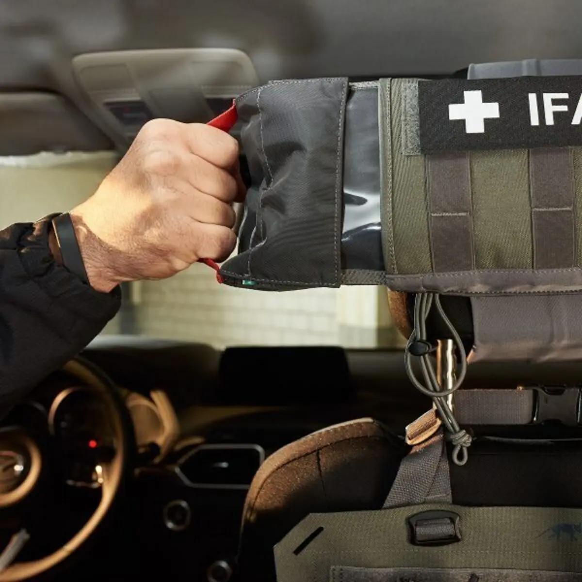 Eine Hand hält das graue TT Head Rest IFAK Erste-Hilfe-Set mit rotem Griff, aufklappbar mit Klettverschluss und "IFAK" Patch, an einer Autokopfstütze befestigt.