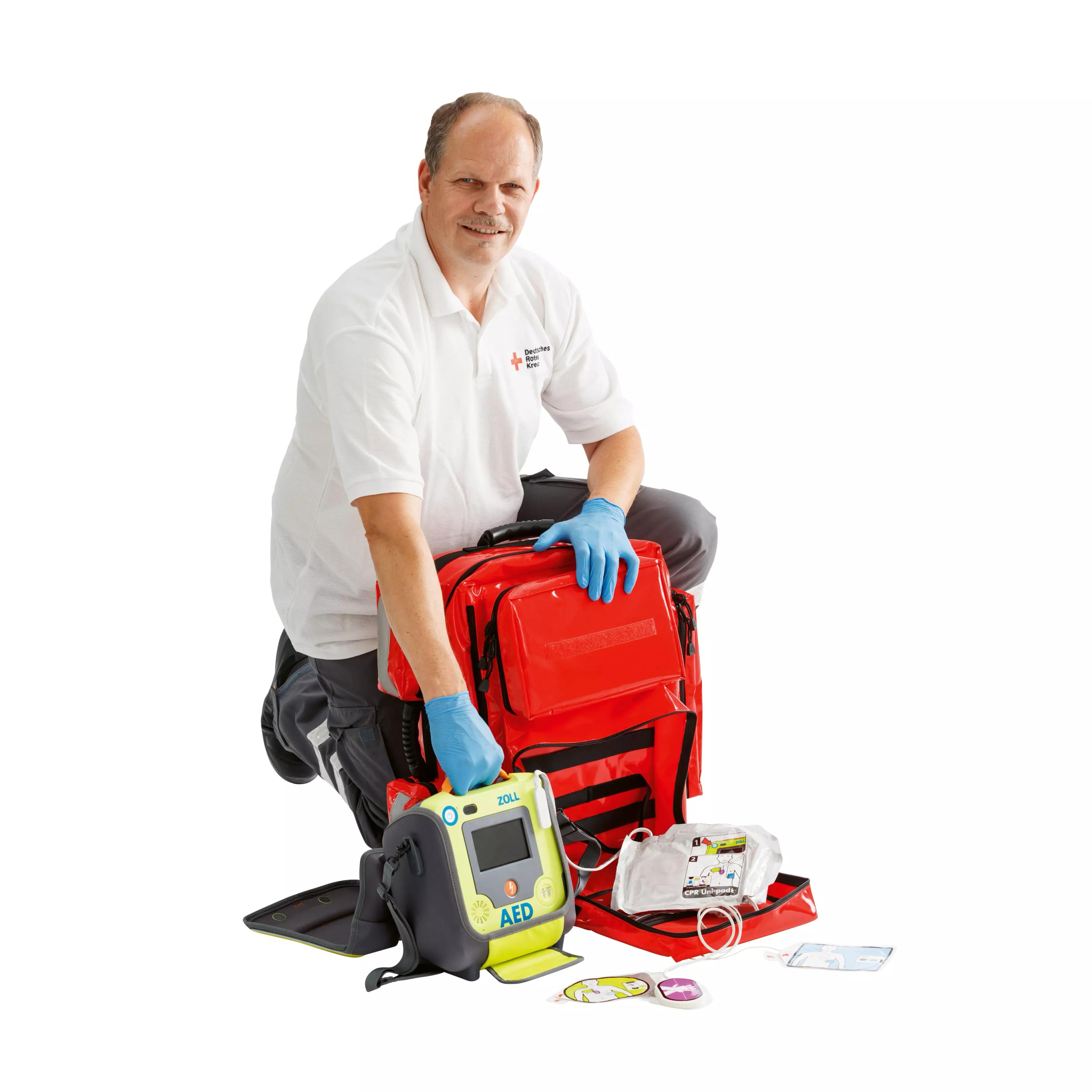 Ein Mann kniet mit blauen Handschuhen und hält einen Übungsdefibrillator ZOLL® AED Plus Trainer2 sowie einen roten Notfallrucksack für Trainingszwecke.
