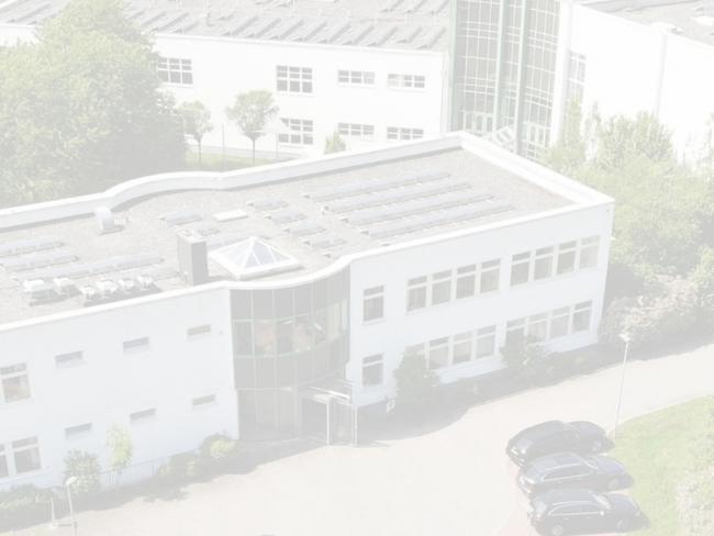 Mehrstöckiges weißes Gebäude mit rechteckigen Fenstern, Solarpanels auf Flachdach, drei Autos auf Parkplatz, umgeben von Bäumen.
