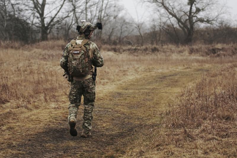 Ein Soldat in Tarnuniform und Helm mit Kamera geht bewaffnet mit Rucksack einen Feldweg entlang durch trockenes Gras.