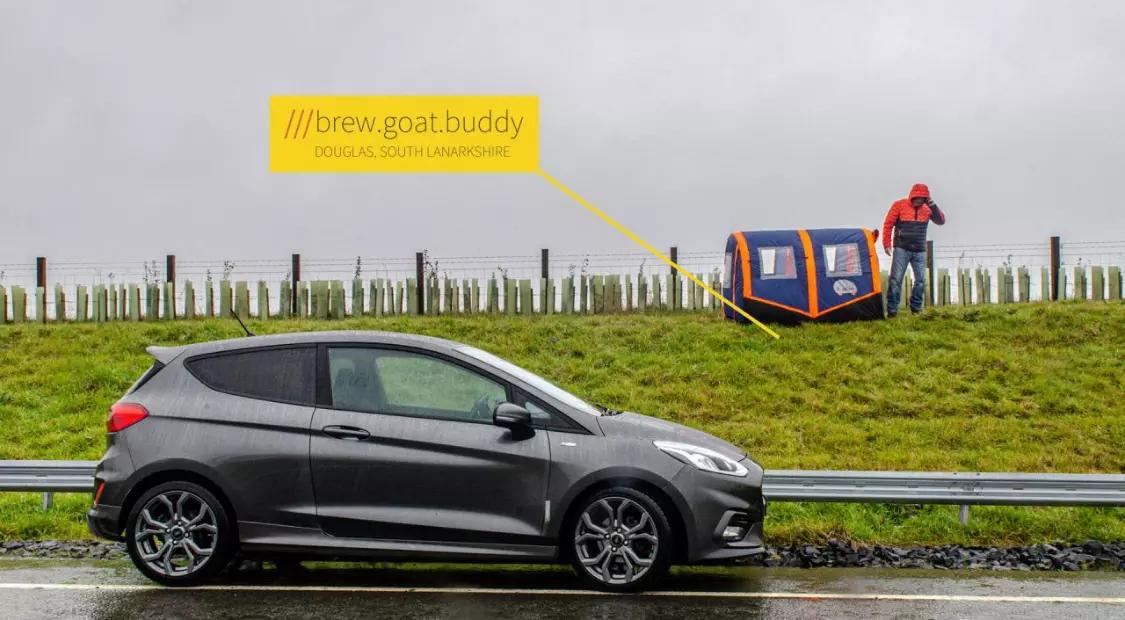 Ein RIGLOO Refuge Notfallzelt in Blau-Orange steht aufgebaut auf feuchtem Gras, nahe einer Person mit Kamera neben einem grauen Ford Fiesta.