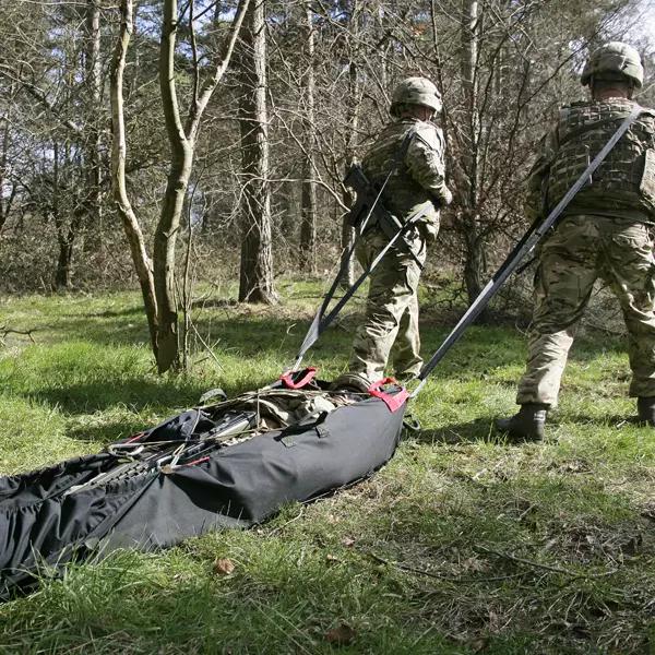Zwei Soldaten ziehen mit den Xtract™ SR Xstraps einen schwarzen Evakuierungsstretcher mit roten Gurten durch bewaldetes Gelände.