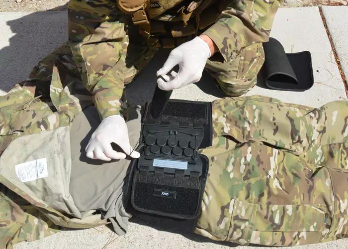 Ein Arrow® T-POD™ - Schwarz wird an einem Bein einer Person mit Tarnhose und weißen Handschuhen befestigt; das ausgeklügelte Seilzugsystem ist sichtbar.