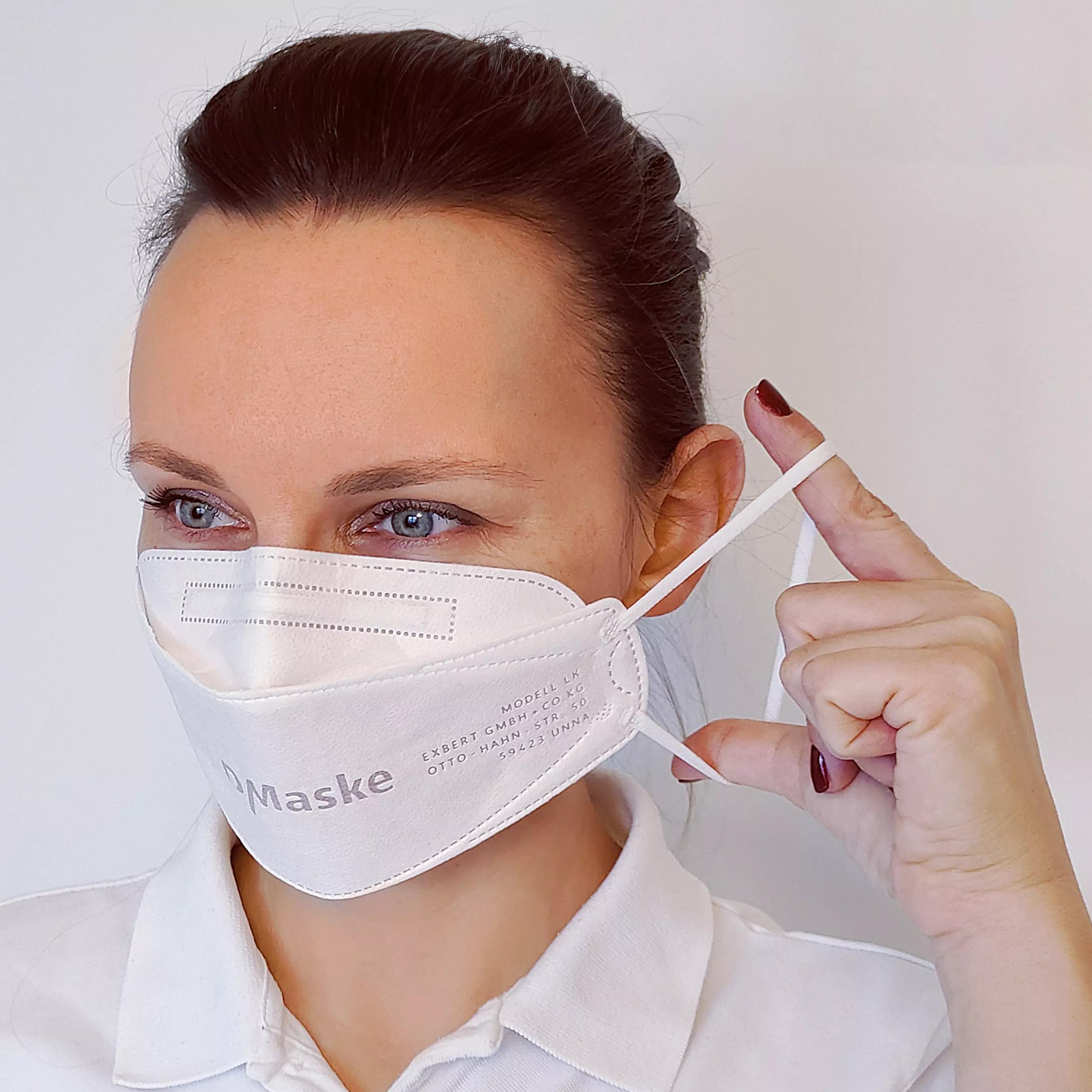 Eine Frau setzt eine Atemschutzmaske FFP2 - MADE IN GERMANY auf, die auffällige schwarze Aufschrift trägt und mit elastischem Band am Ohr befestigt wird.