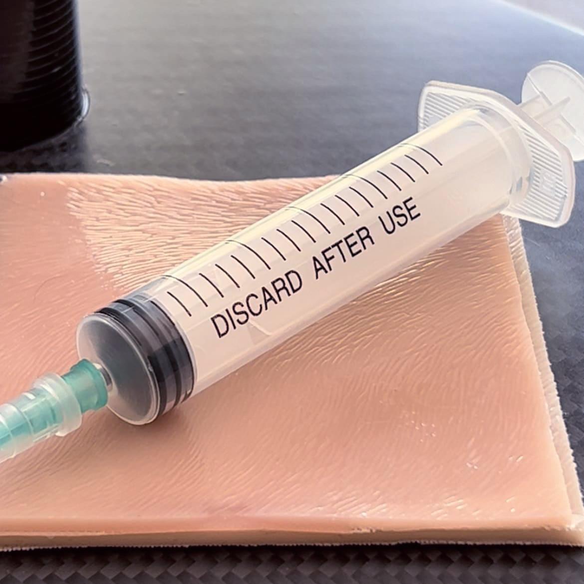 Eine transparente Spritze mit schwarzem Kolben und grün-verschlossener Nadel liegt neben einem Battlefield Partners Ersatzstreifen für den Tragbaren CRIC Trainer.