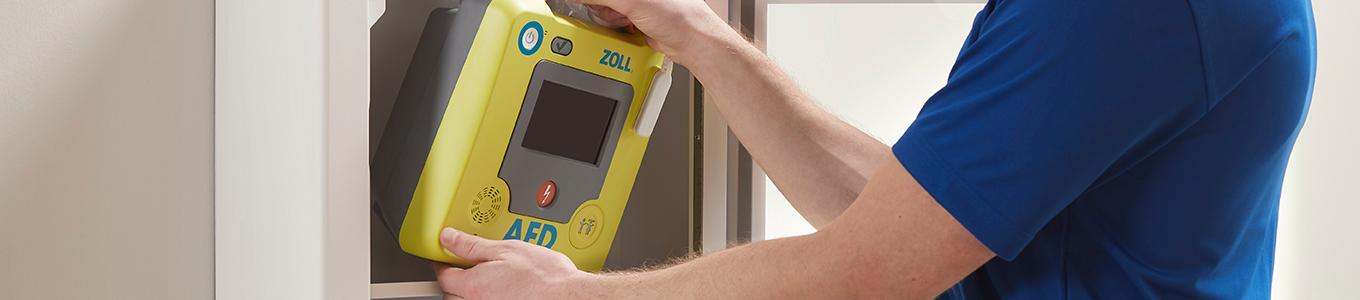 Eine Person bedient einen gelb-grauen Defibrillator mit Display und rotem Notfallknopf, der an einer weißen Wand montiert ist.
