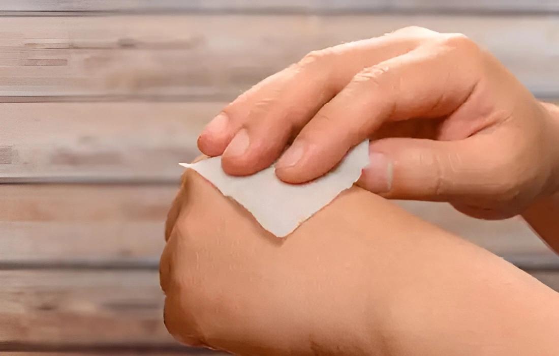 Hand hält weißen Wattepad und reinigt damit sanft die Haut vor einer hellen Holzfläche mit waagerechten Planken.