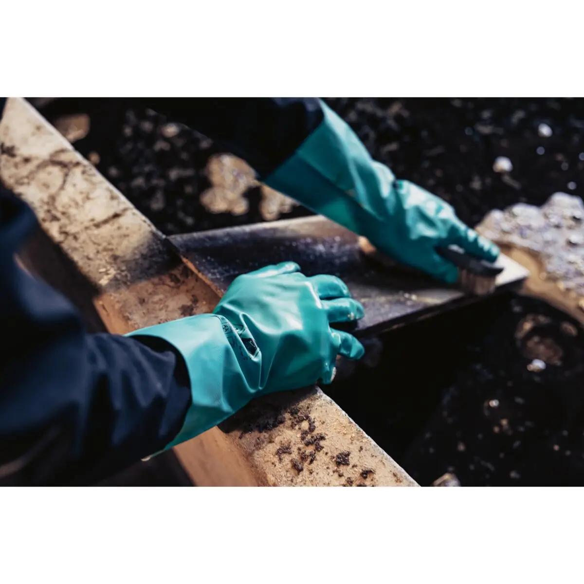 Zwei Hände tragen Chemikalienschutzhandschuh Nitril 33, grün, beim Reinigen einer verschmutzten Metallfläche mit Bürste im rechteckigen Behälter.