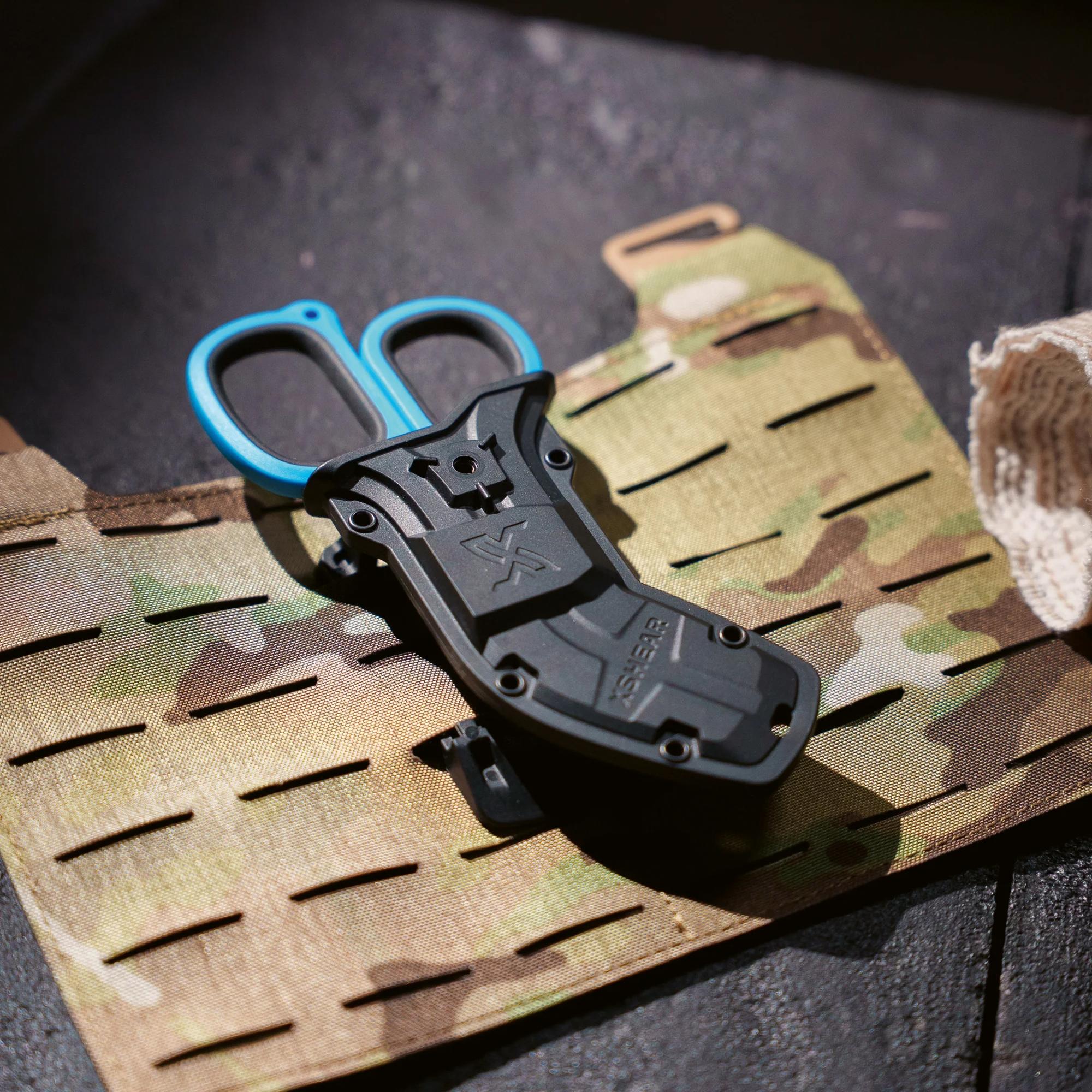 Das XSHEAR® Mini Tactical Holster hält eine blaue Schere sicher an einer tarnfarbenen taktischen Weste mit horizontalen Schlitzen und MOLLE-kompatiblem Gürtelclip.