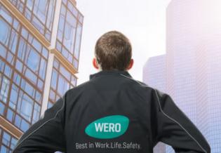 Person in schwarzer Jacke mit türkisfarbenem WERO-Logo und weißem Ärmelabschluss steht vor einem modernen Hochhaus mit Glasfassade.