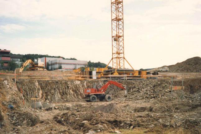 Orangefarbener Bagger hebt Erde auf Baustelle, gelber Kran im Hintergrund, mehrstöckiges Gebäude im Rohbau, roter Walzenfahrzeug daneben.