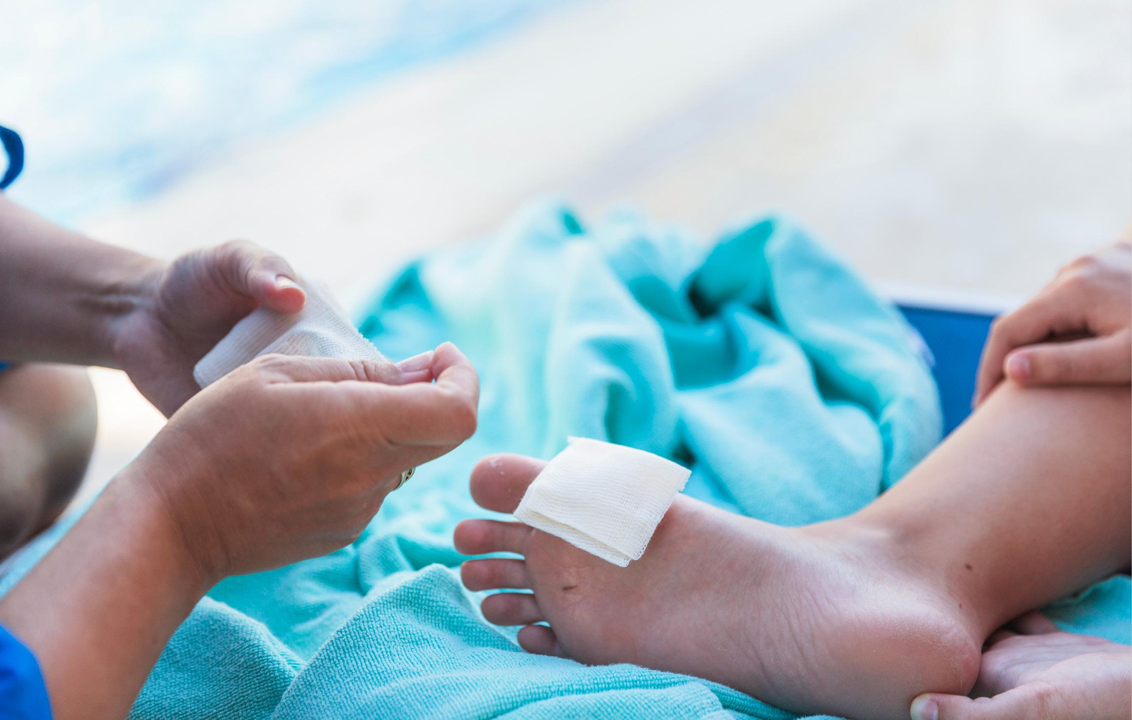 Ein Kind hat sich am Fuß verletzt und wird auf einem Handtuch sitzend mit einem Pflaster versorgt., Hand, Person, Baby, Finger, Massieren