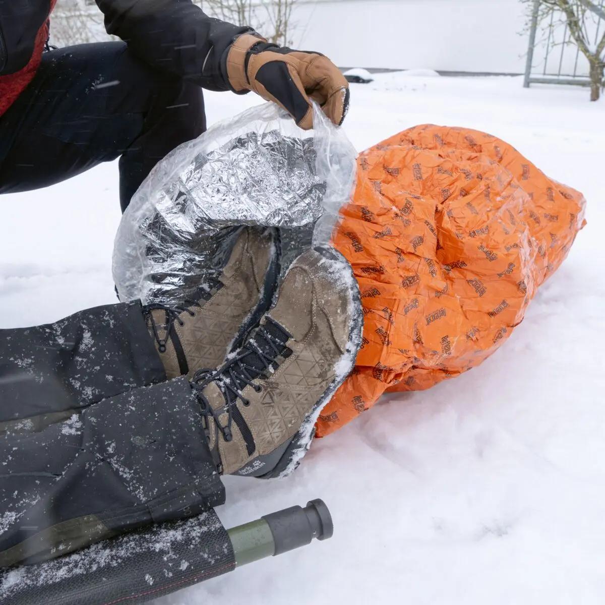 Ein Blizzard - Orange, Schlafsack mit Reflexcell™ Mehrlagen-Isolierung liegt im Schnee, neben winterfesten Stiefeln und Campingausrüstung.