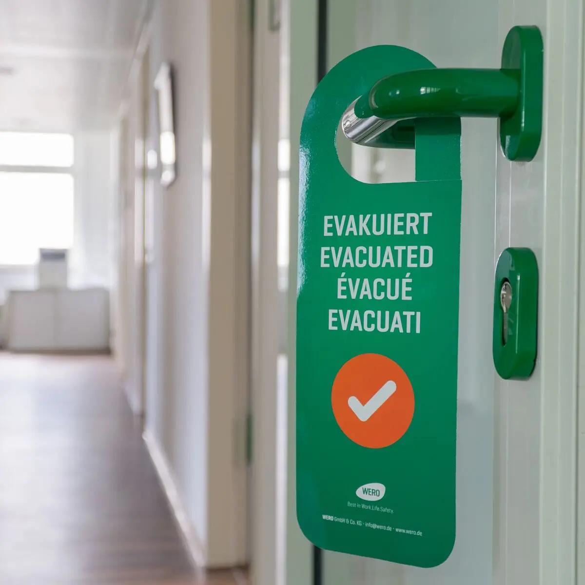 Grüner Türanhänger "Evakuiert" mit weißer Aufschrift, rotem Haken und beidseitigem Hinweis an einer Glastür im Flur sichtbar angebracht.