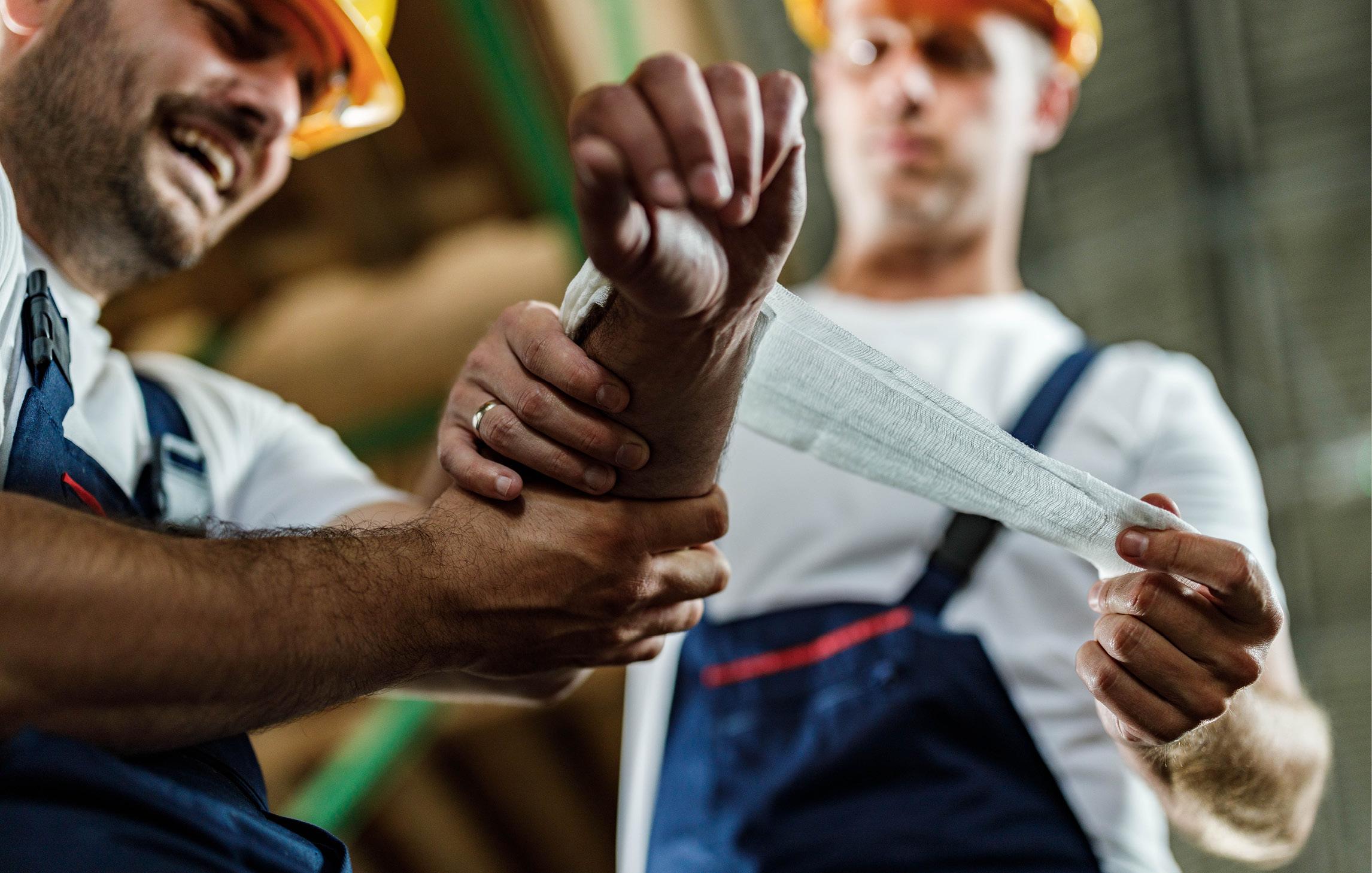 Ein Arbeiter hat sich am Arm verletzt und wird von einem Kollegen versorgt. Dieser wickelt eine Mullbinde um den Arm