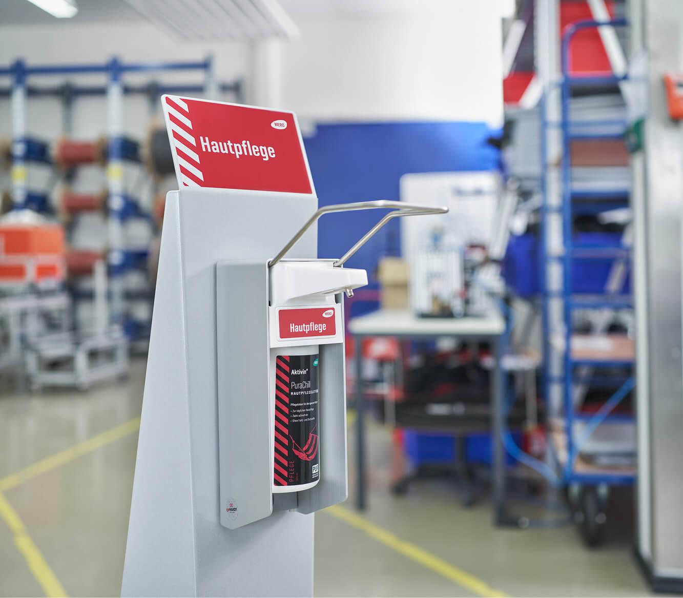 Weiß-roter Activit Handhygienespender mit silberner Pumpe und rotem "Hautpflege" Schild steht in einer Werkstatt.