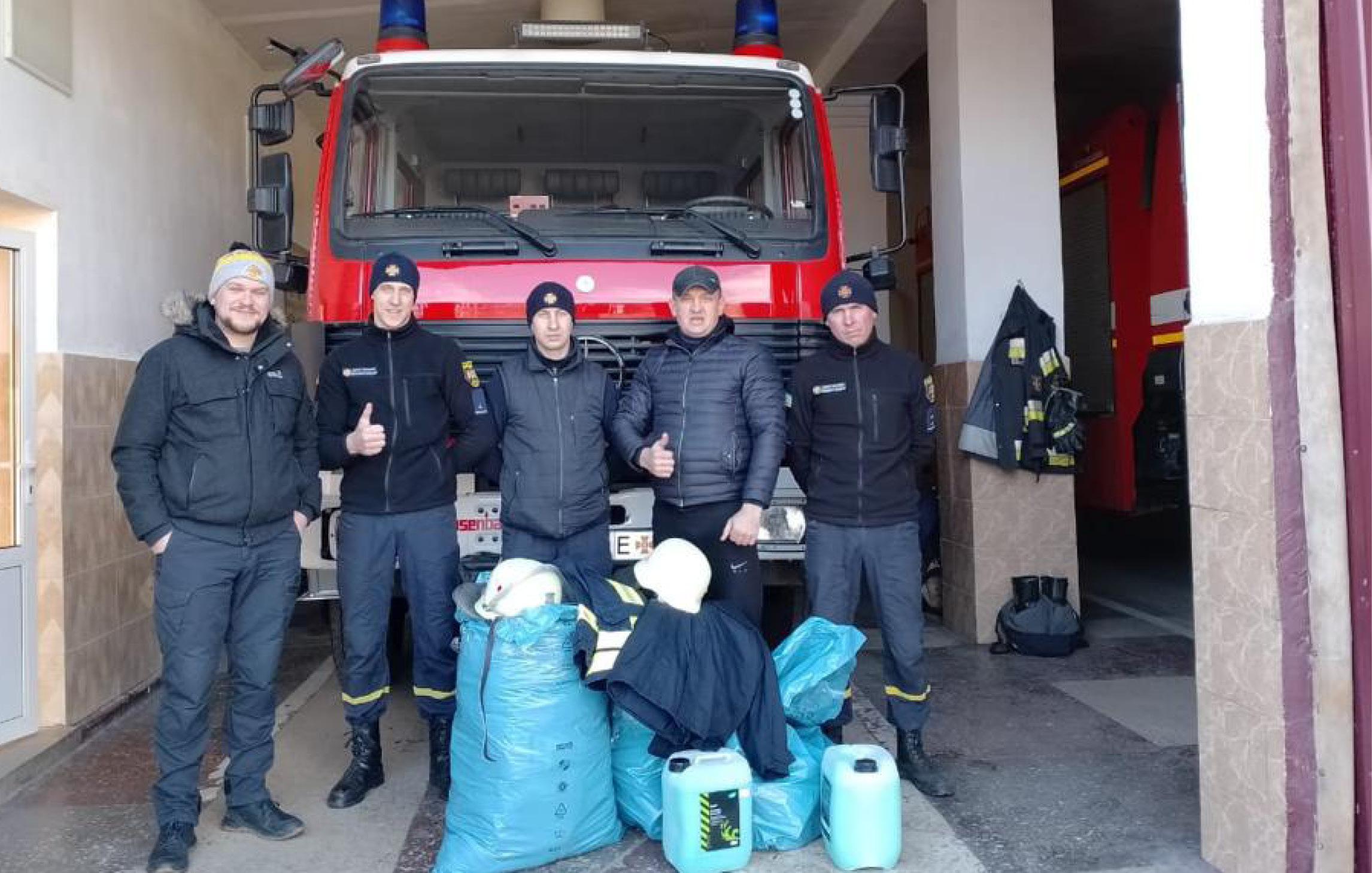 Sechs Feuerwehrleute in Uniform stehen vor einem roten Feuerwehrfahrzeug, zwei halten große Ausrüstungs- oder Materialtaschen.