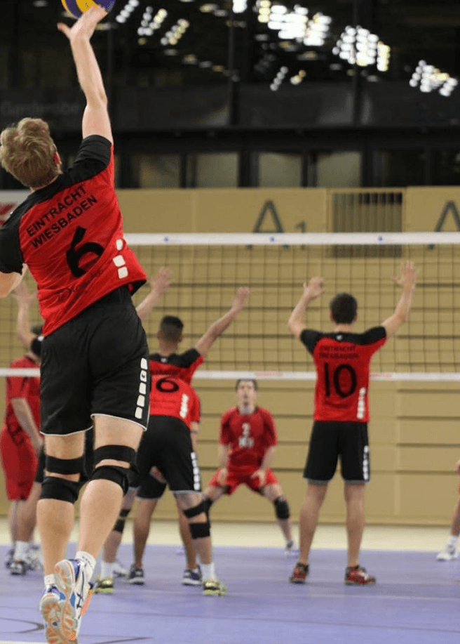 Mehrere Volleyballspieler in roten und schwarzen Trikots spielen während ein Spieler am Netz zum Sprungschlag ansetzt auf einem lila Hallenfeld.