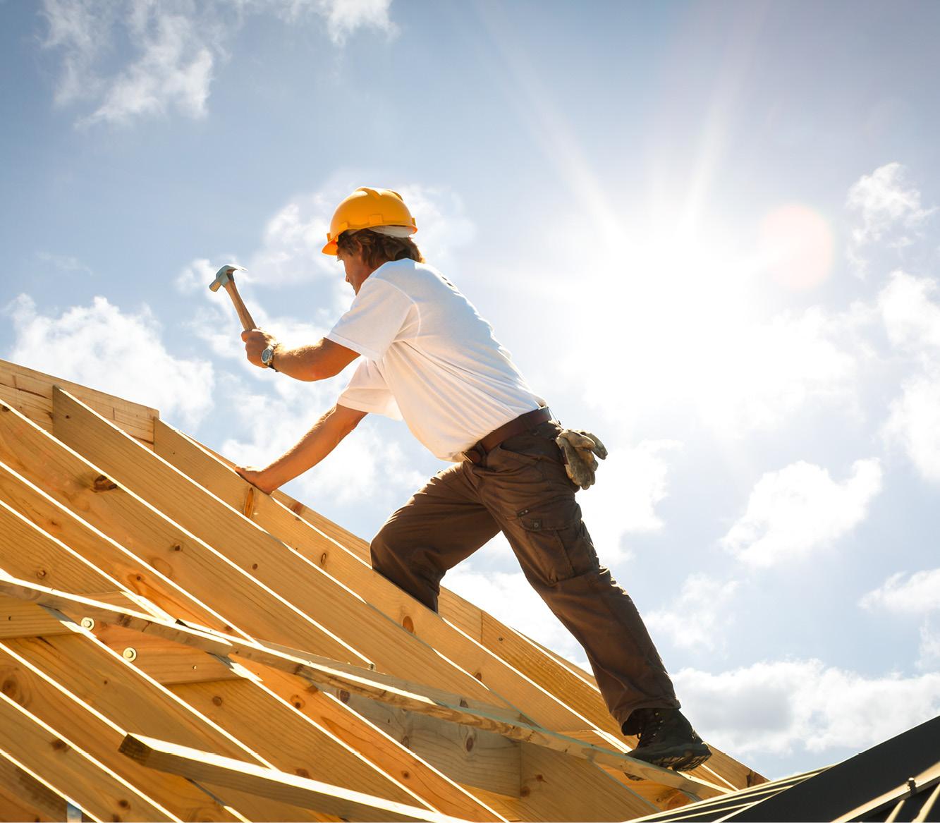 Ein Bauarbeiter mit gelbem Helm befestigt Holzbalken mit einem Hammer auf einem teilfertigen Dachstuhl aus Holzträgern.