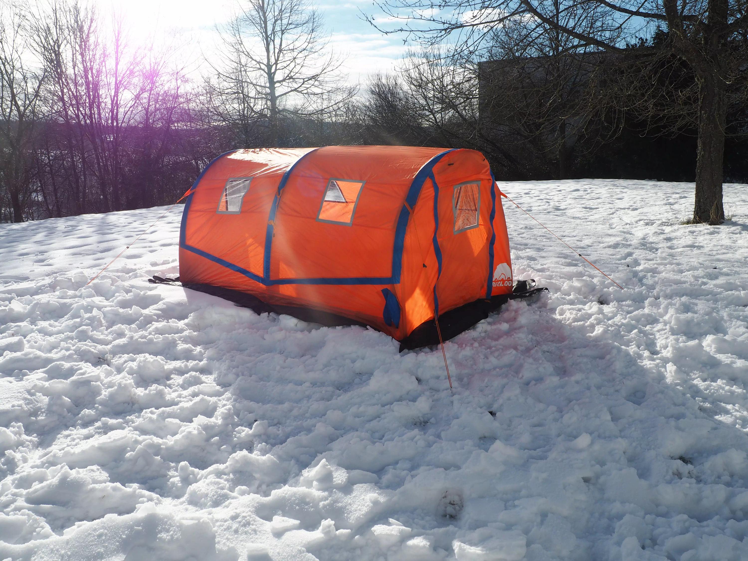 Ein orange-blaues RIGLOO Rescuer Rettungszelt mit zwei Fenstern und zwei Gestängebögen steht einsam auf verschneitem Feld, umliegend Bäume.