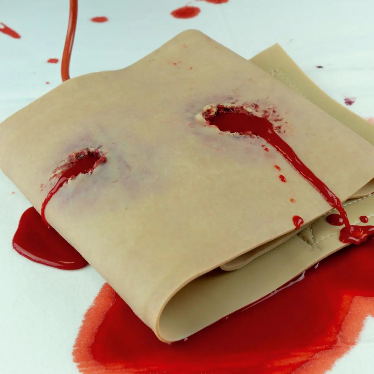 Eine Techline Moulage Schusswunde Bein Simulationstuch mit mehreren durchgehenden Schusswunden und sichtbarem Kunstblut auf weißer Fläche.