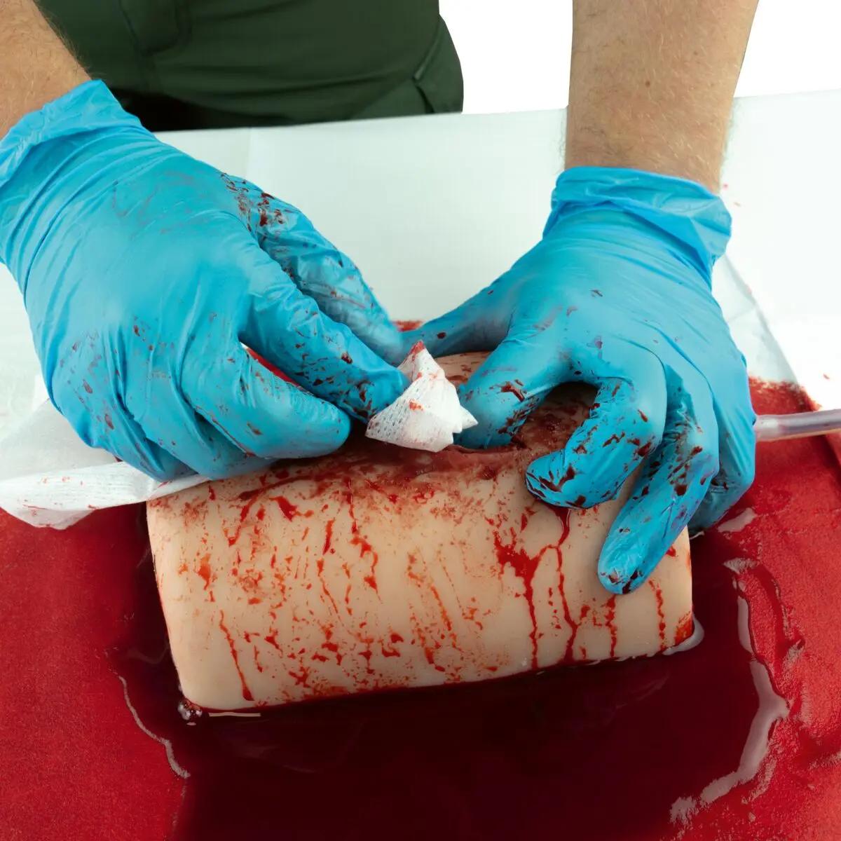 Zwei behandschuhte Hände reinigen das TrueClot® Wound Packing Trainingssystem Schnittwunde, das mit realistischem Kunstblut für Blutstillungstraining bedeckt ist.