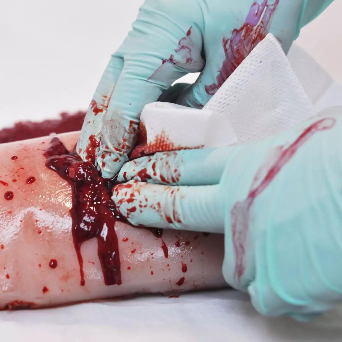 Zwei behandschuhte Hände reinigen mit Tuch und Bürste eine blutverschmierte Wunde am TrueClot® Woundpacking Trainingssystem - Schusswunde, um realistisches Blutstillungstraining zu demonstrieren.