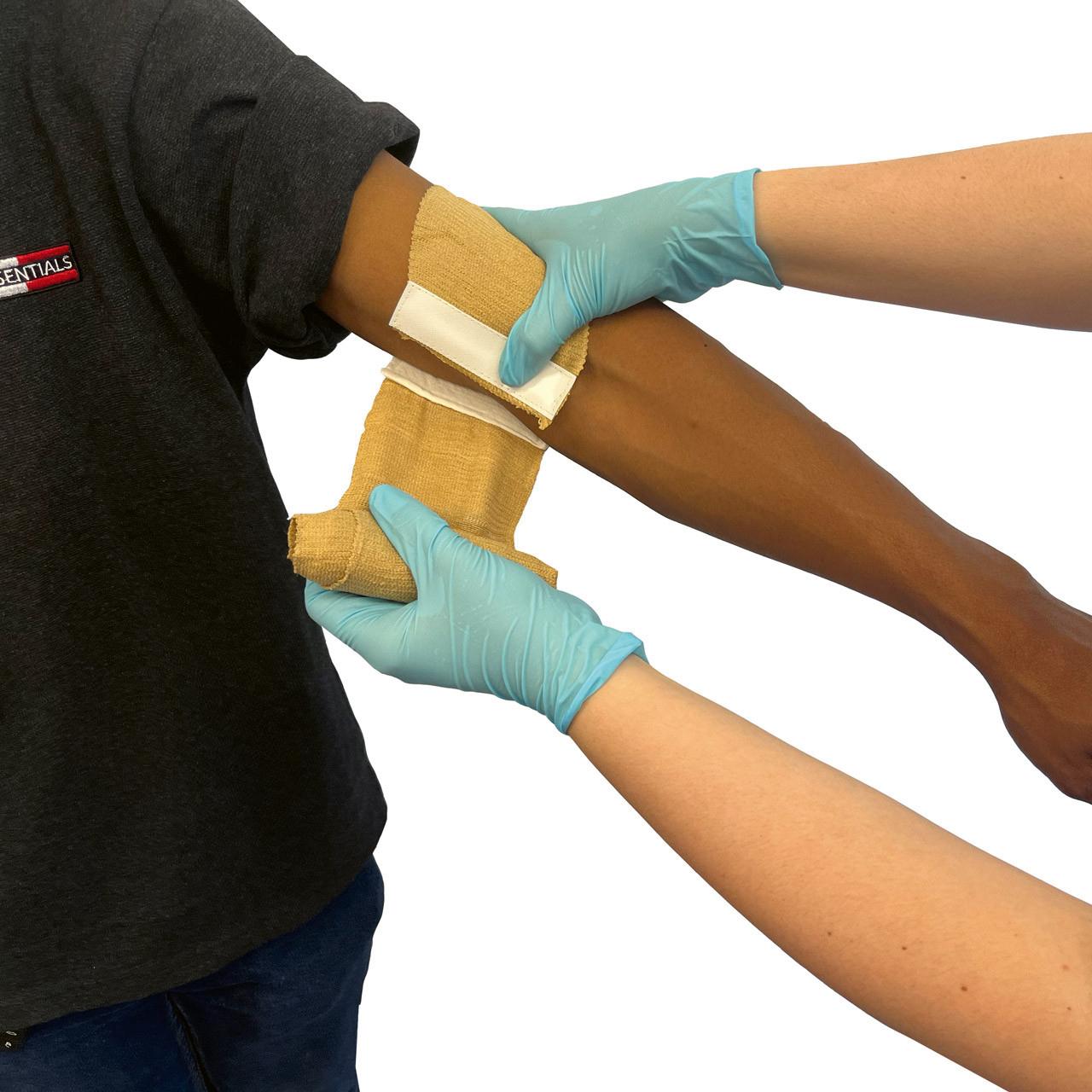 Eine Rescue Essentials® Mini Compression Bandage wird von zwei behandschuhten Händen fest um den ausgestreckten Arm einer dunkelhäutigen Person gewickelt.