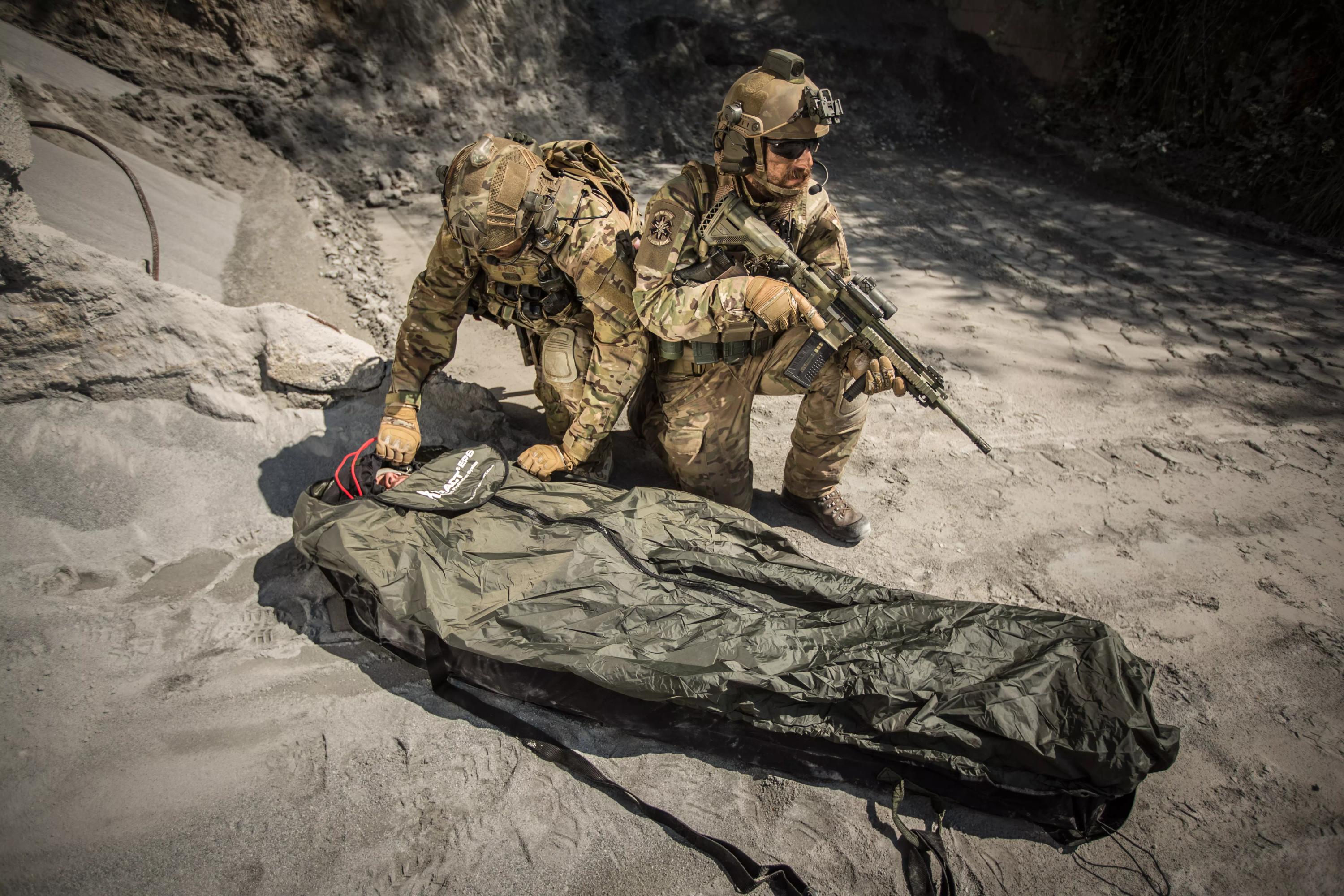 Zwei Soldaten in Tarnuniform verwenden das Xtract™ SR Element Protection System (EPS), um eine Person mit einem grünen Schutzüberzug im felsigen Gelände vor Unterkühlung zu schützen.