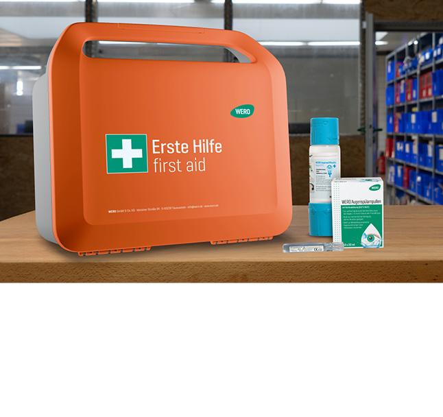 Oranger Erste-Hilfe-Kasten mit weißem Kreuz, daneben blaue Flasche, WERO-Box und transparenter Schlauch auf Holztisch