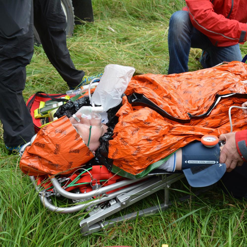 Eine Person mit Blizzard - Orange, Beanie liegt auf einer Trage im Freien, ihr Bein ruht erhöht, ein Beatmungsschlauch und eine Infusion sind gelegt.