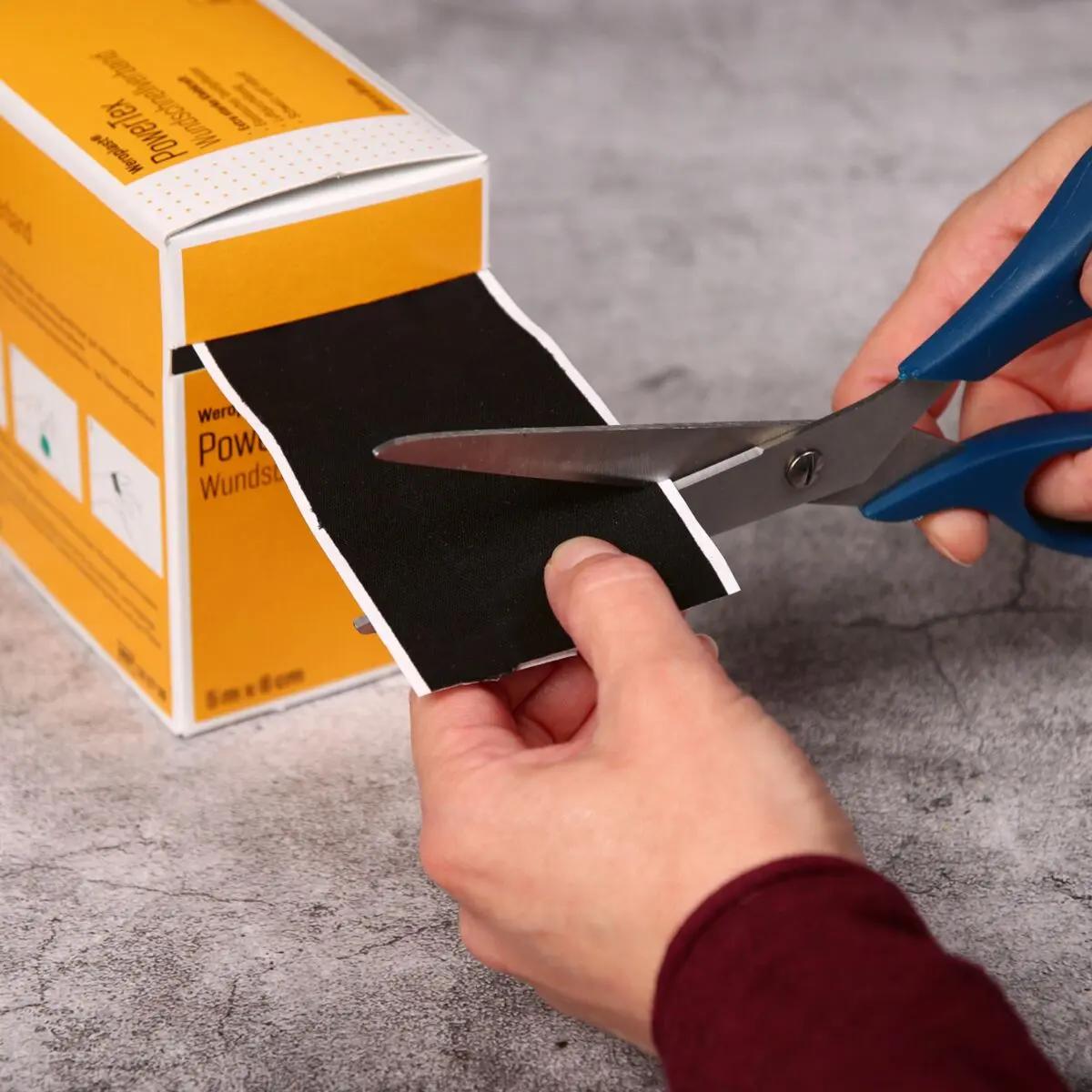 Eine Hand hält eine Schere mit blauen Griffen und schneidet gerade ein Stück des Wundschnellverband Weroplast® PowerTex von einer Verpackung ab.