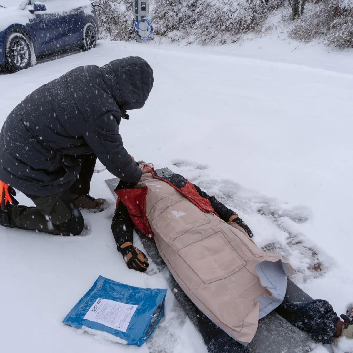 Eine Person legt im Schnee einem anderen eine beige Ready-Heat™ - 6-Element-Decke an, die zur schnellen Erwärmung und zum Kälteschutz dient.