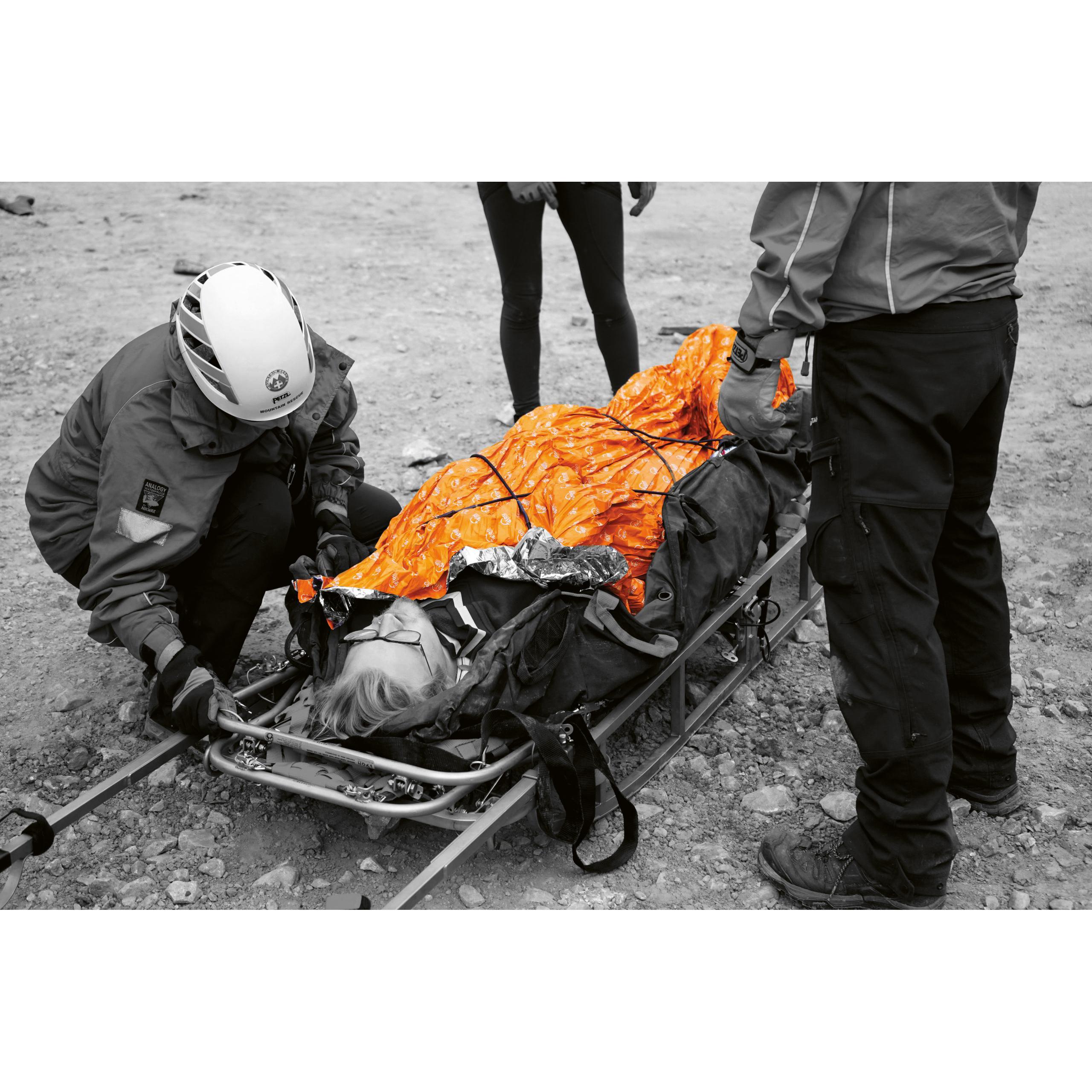 Eine Person liegt auf einer Trage und ist komplett in eine igloo Survival Pro - Orange, Größe L Decke gehüllt, während Helfer daneben knien.