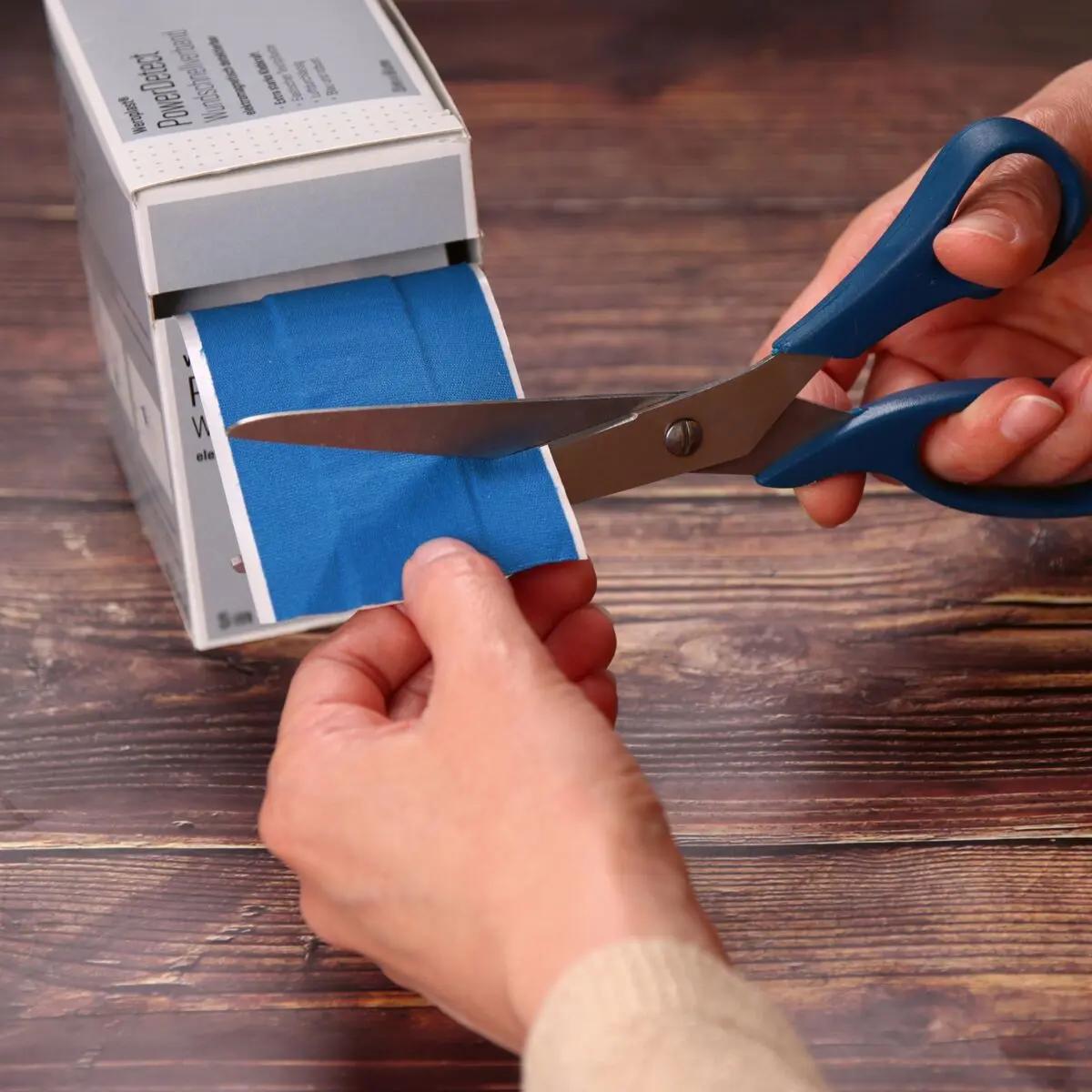 Eine Hand schneidet mit einer blauen Schere eine Bahn des Wundschnellverband Weroplast® PowerDetect aus blauem, detektierbarem, elastischem Textilgewebe ab.