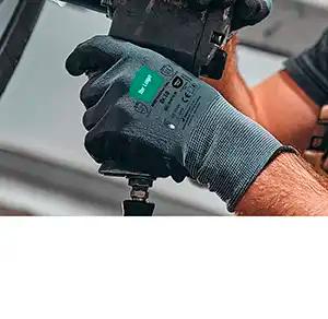 Eine Hand mit dunkelblauem Arbeitshandschuh hält ein schwarzes Elektrowerkzeug mit grünem Etikett senkrecht am Griff.