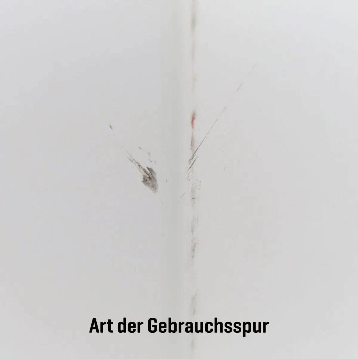 Weiße Wand mit zwei schwarzen Schrammen und Rissen als Beispiel für Gebrauchsspuren am AED-Wandschrank (gebraucht) - mit Alarmfunktion (Sirene & Leuchte).