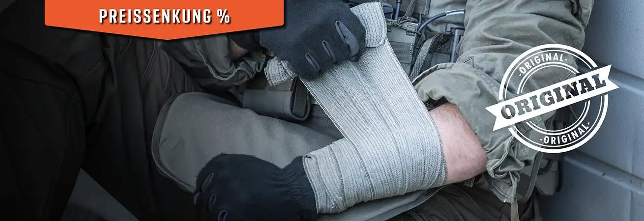 Eine Person mit dunklen Handschuhen wickelt eine elastische Bandage um ihren Unterarm; sie trägt eine taktische Weste.