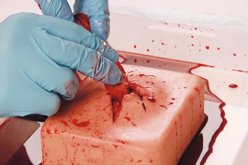 Eine Hand mit blauen Handschuhen führt eine Wundversorgung an einem Erler-Zimmer Wound Packing Trainer mit realistischer Schusswunde und Blutungsfunktion durch.