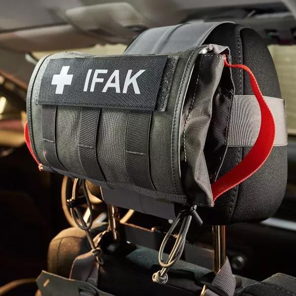 Schwarzes IFAK Erste-Hilfe-Set mit weißem Logo, per rotem Gurt an einer schwarzen Auto-Kopfstütze befestigt, mehrere Fächer sichtbar.
