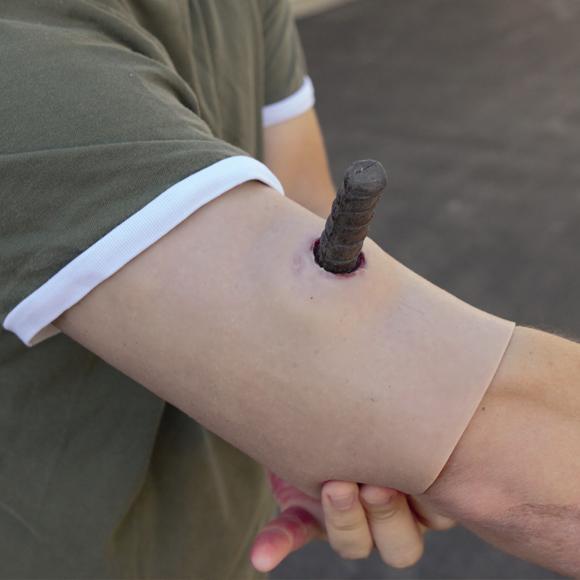 Techline Moulage Aufgespießtes Objekt zeigt einen angewinkelten Arm mit einem dunklen, stählernen Bolzen, der realistisch aus der Haut hervorragt.