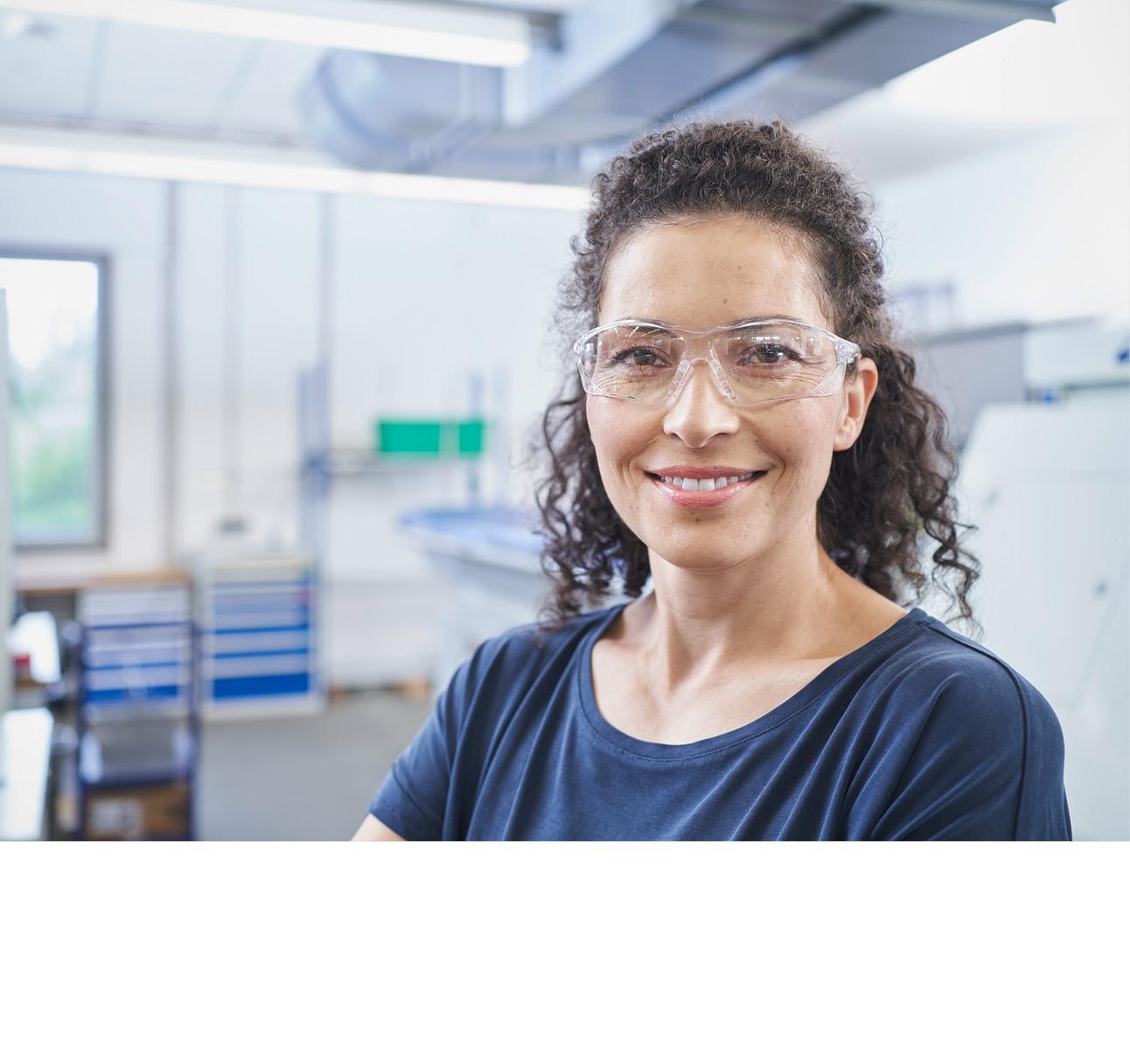 Frau mit farbloser Schutzbrille steht in einer Werkstatt