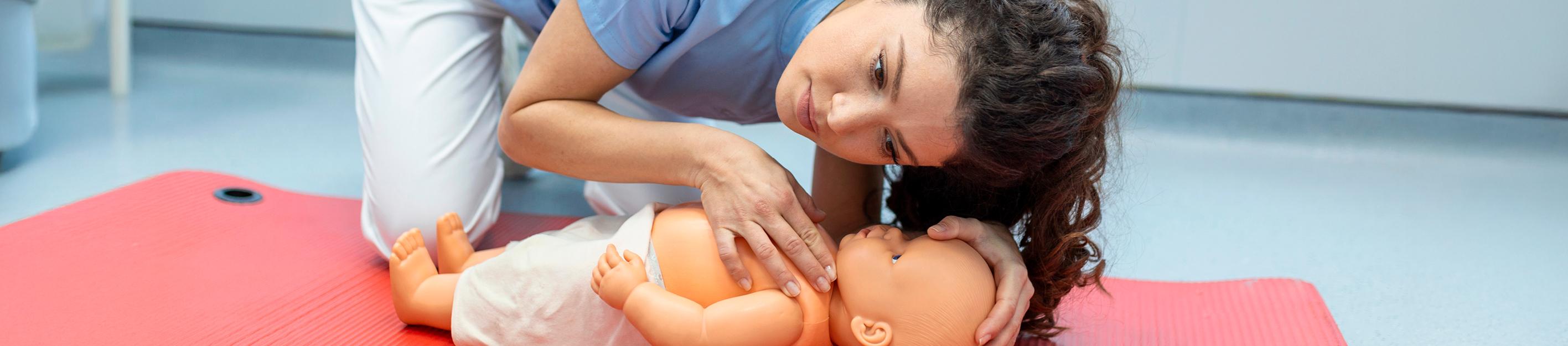 Eine Frau in blauer Kasack und weißer Hose übt an einem Babypuppen-Torso auf einer roten Matte die Herzdruckmassage.