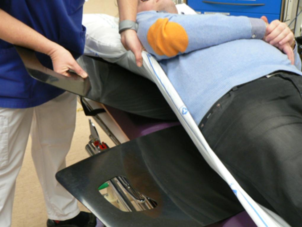 Eine Person im hellblauen Pullover liegt auf einer Trage, während ein medizinischer Fachmann sie mit einer metallenen Fläche unterstützt.