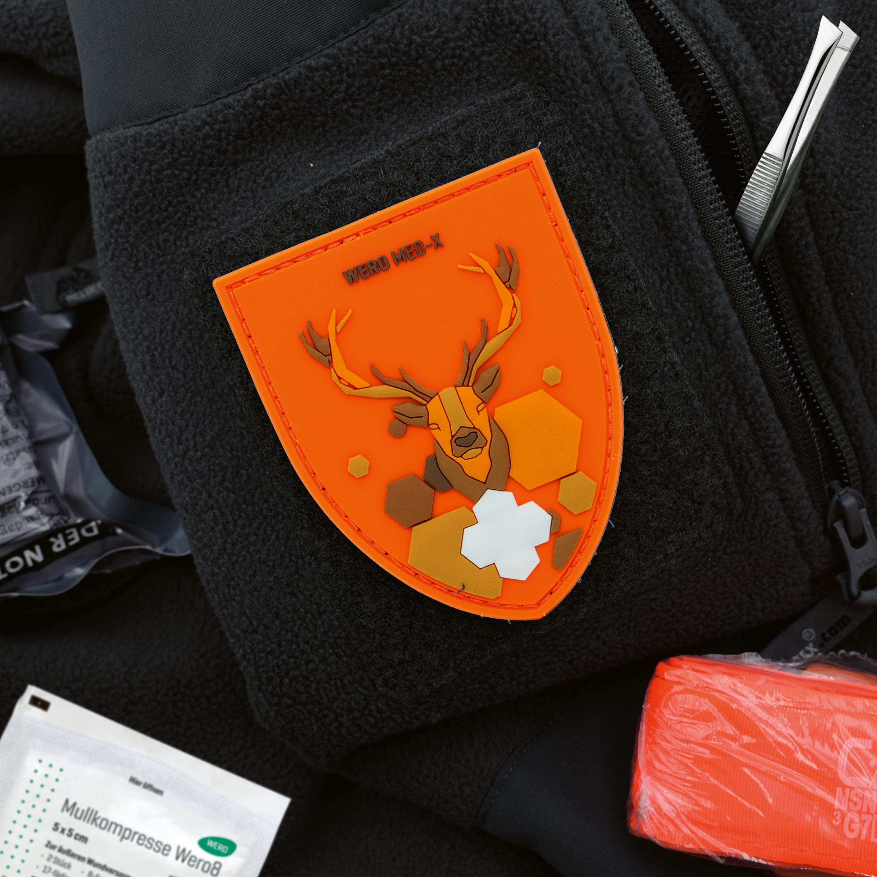 Ein Patch WERO MED-X® Hunter Medic Pack mit stilisiertem Hirschkopf auf schwarzer Fleecejacke, daneben Erste-Hilfe-Zubehör wie Verbandpäckchen und Pinzette.