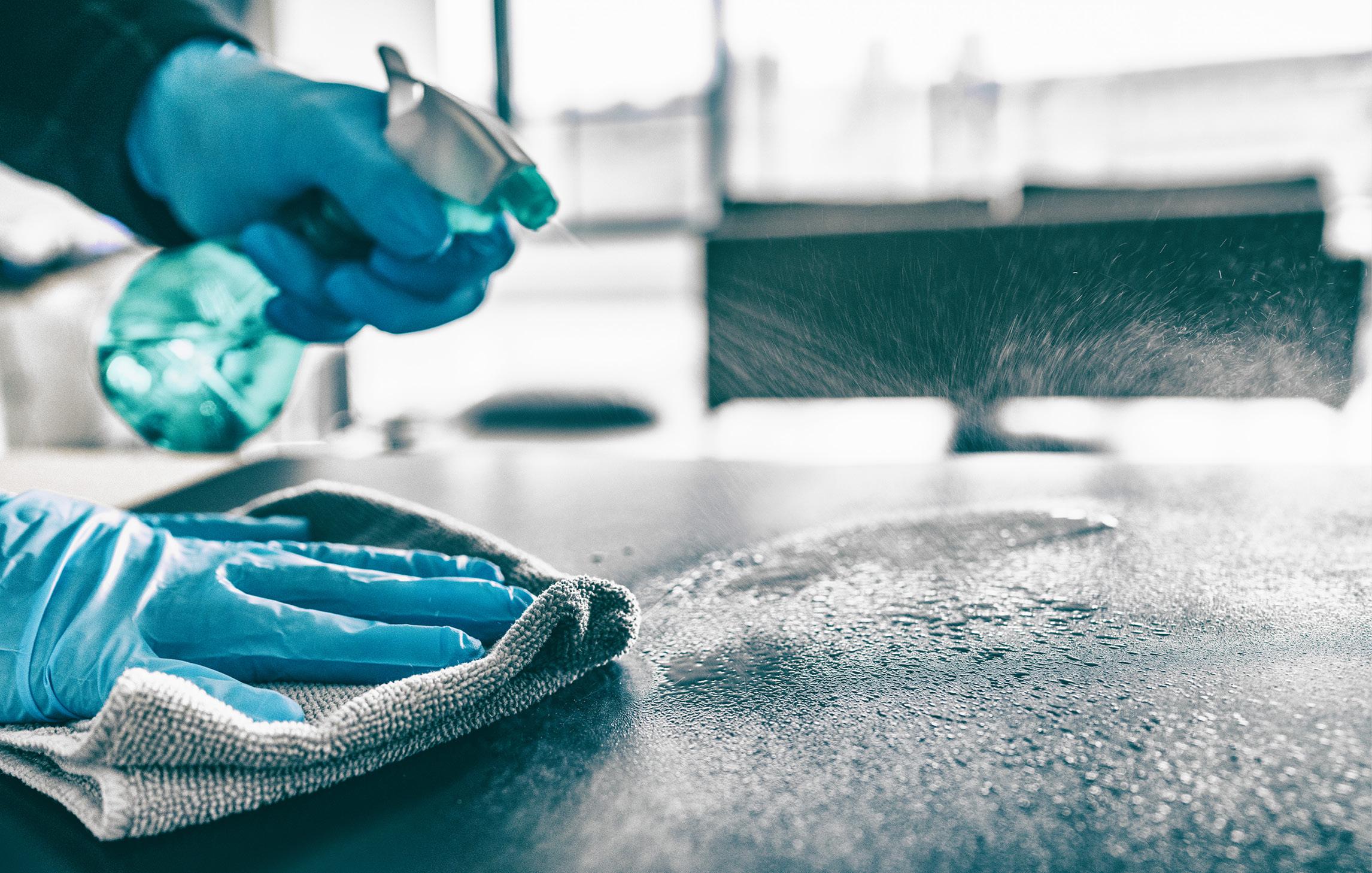 Eine Oberfläche wird desinfiziert. Die Person trägt blaue Einmalhandschuhe, einen Stofflappen und eine Sprühflasche