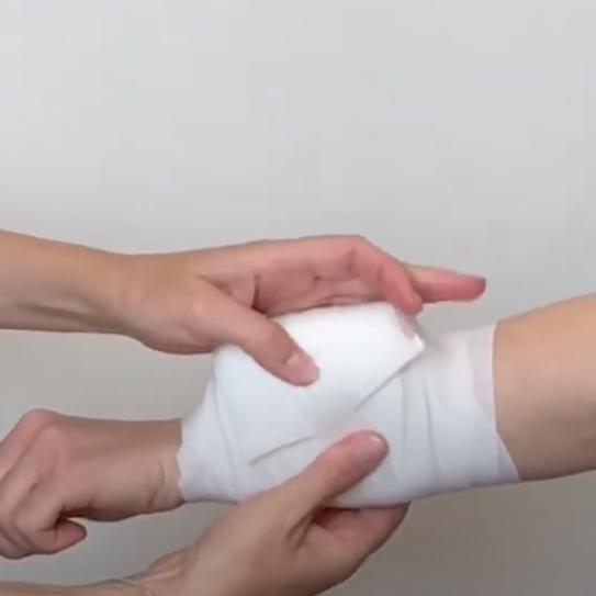 Ein gebogener Arm mit heller Haut wird mit weißen Mullbinden verbunden, die von einer Hand zum Fixieren sanft gehalten werden.