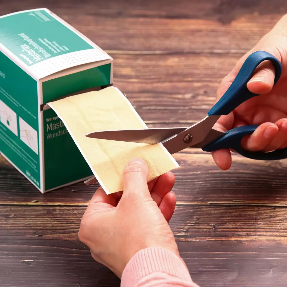 Eine Hand schneidet mit einer blauen Schere ein Stück des Wundschnellverband Weroplast® MasterTex aus gelblichem, anpassungsfähigem Gewebe von der Rolle ab.