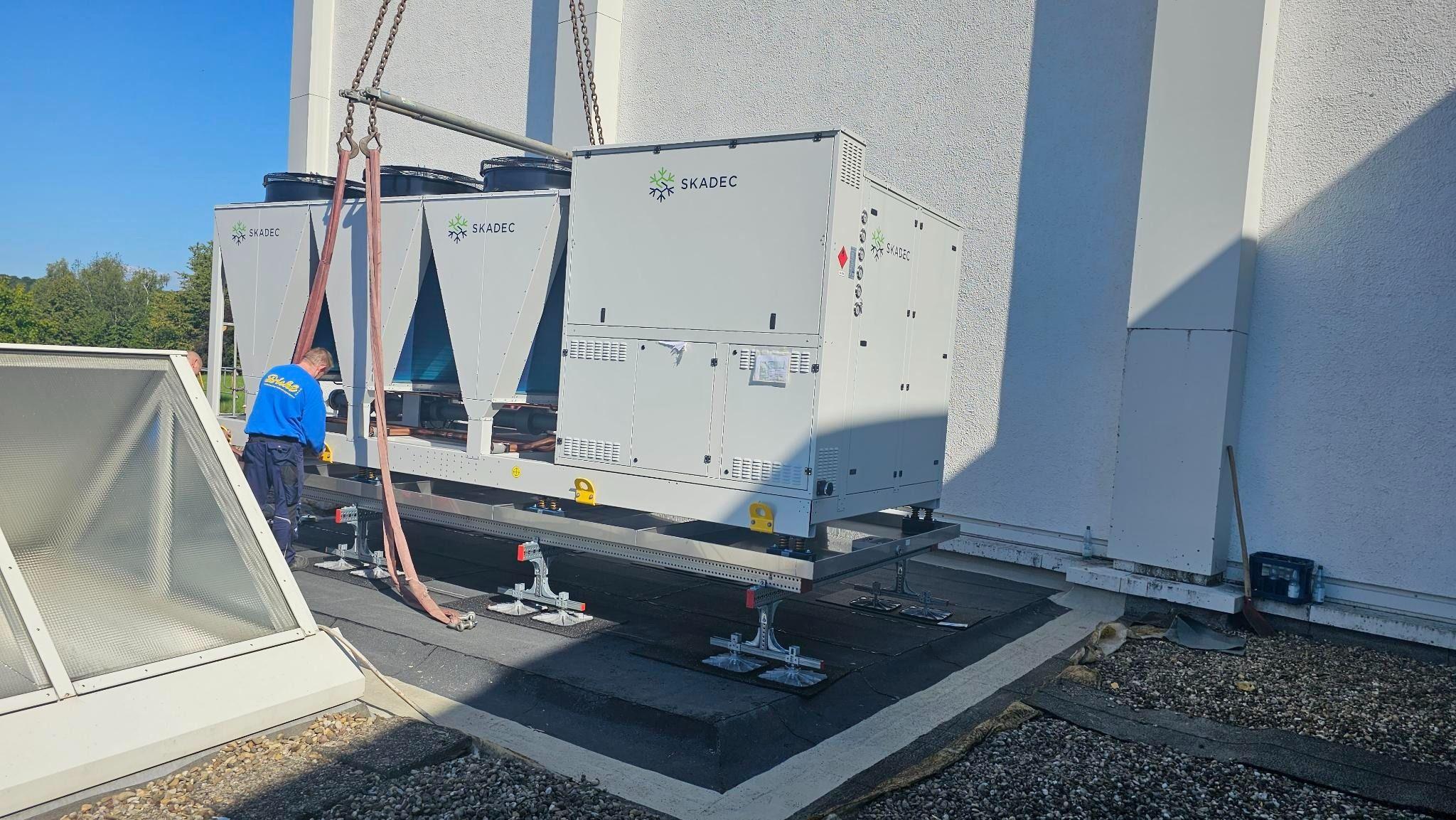 Große Skadec Klimaanlage mit vier Ventilatoren wird mithilfe von Ketten auf das Dach eines weißen Gebäudes montiert, Arbeiter assistiert.